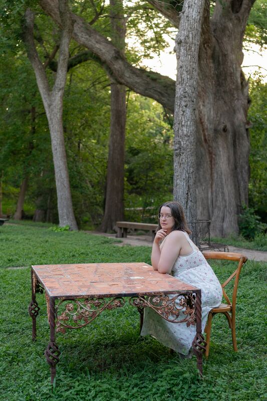 Woman seated at a wrought-iron tile table under tall trees